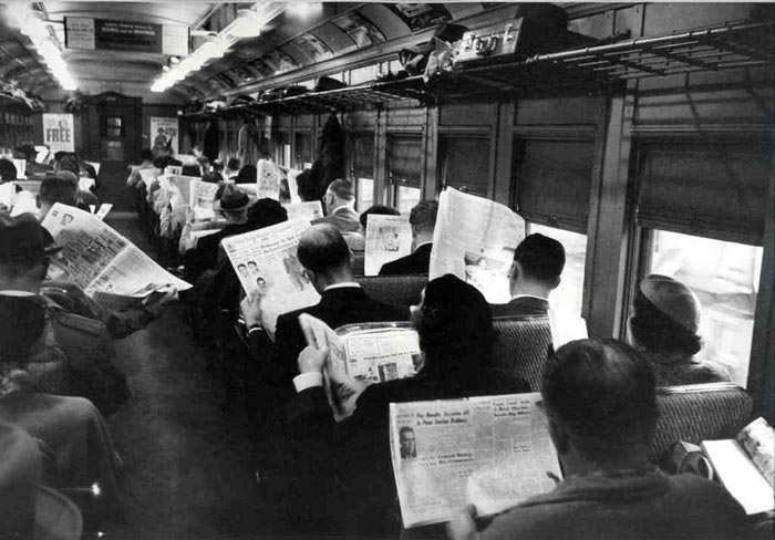 A mid-20th century train carriage full of passengers reading news papers